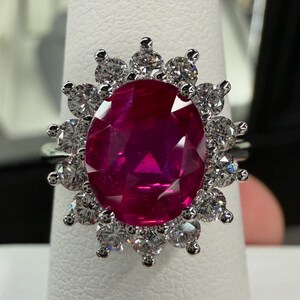 May include: A silver-toned ring featuring a large, oval-cut, deep pinkish-red gemstone at the center. The central stone is surrounded by a halo of twelve small, round, clear, faceted stones set in a pronged arrangement on a metallic band.