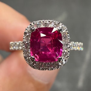May include: A ring featuring a large, cushion-cut, vibrant pink gemstone set in a silver-toned metal band. The central stone is surrounded by a halo of small, round, clear, sparkling stones, with additional small stones set along the band.