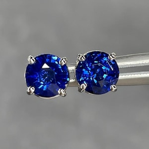 Pode incluir: Um par de brincos de bot&atilde;o de safira azul redondos e brilhantes. Cada pedra preciosa &eacute; colocada em uma configura&ccedil;&atilde;o de prata de quatro pontas. As safiras exibem uma cor azul profunda e vibrante e s&atilde;o presas por uma ferramenta de prata em um fundo cinza.