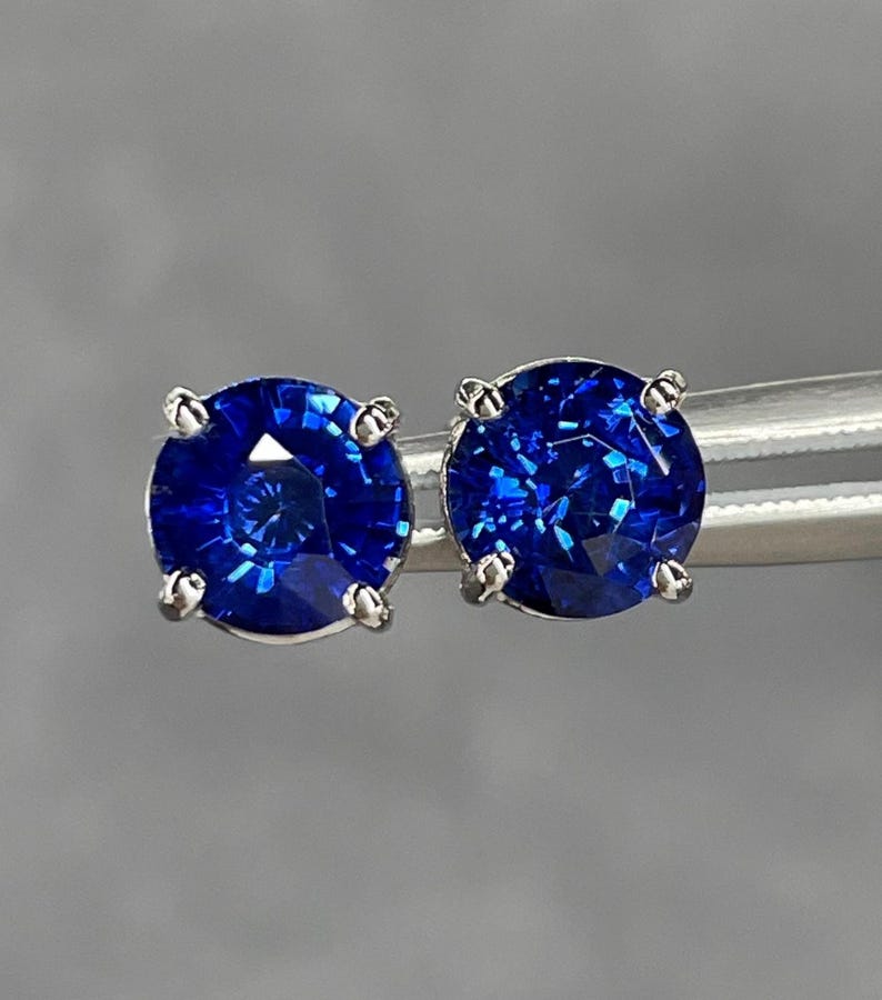 Pode incluir: Um par de brincos de bot&atilde;o de safira azul redondos e brilhantes. Cada pedra preciosa &eacute; colocada em uma configura&ccedil;&atilde;o de prata de quatro pontas. As safiras exibem uma cor azul profunda e vibrante e s&atilde;o presas por uma ferramenta de prata em um fundo cinza.