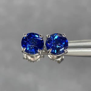 Pode incluir: Um par de brincos de press&atilde;o com pedras preciosas redondas, azuis escuras. As pedras s&atilde;o facetadas e engastadas em metal prateado com quatro garras. Os brincos s&atilde;o exibidos em uma superf&iacute;cie reflexiva.