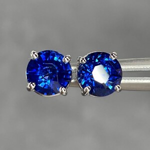 Pode incluir: Um par de brincos de press&atilde;o com pedras preciosas redondas, azuis profundas e brilhantes. As pedras s&atilde;o engastadas em metal prateado com quatro garras. Os brincos s&atilde;o exibidos em um fundo cinza neutro.