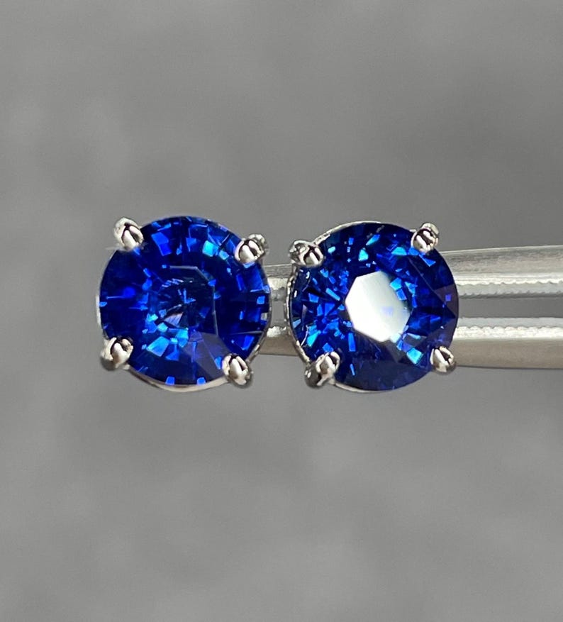 Pode incluir: Um par de brincos de press&atilde;o com pedras preciosas redondas, azuis profundas e brilhantes. As pedras s&atilde;o engastadas em metal prateado com quatro garras. Os brincos s&atilde;o exibidos em um fundo cinza neutro.