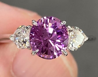 Pode incluir: Um anel de noivado de safira roxa com dois diamantes em forma de coração de cada lado da pedra central.
