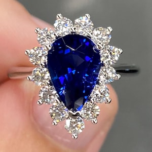 May include: A silver ring featuring a pear-shaped, deep blue gemstone surrounded by a halo of smaller, round, clear gemstones. The ring's design is elegant and eye-catching, suitable for special occasions.