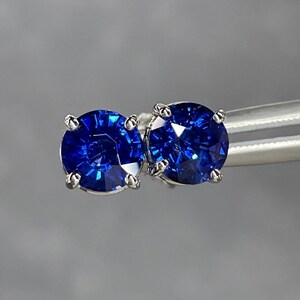 Pode incluir: Um par de brincos de press&atilde;o de safira azul redondos com corte brilhante. Cada brinco apresenta uma gema azul profunda e vibrante em uma crava&ccedil;&atilde;o de garras prateada. Os brincos s&atilde;o exibidos em um fundo cinza suave.