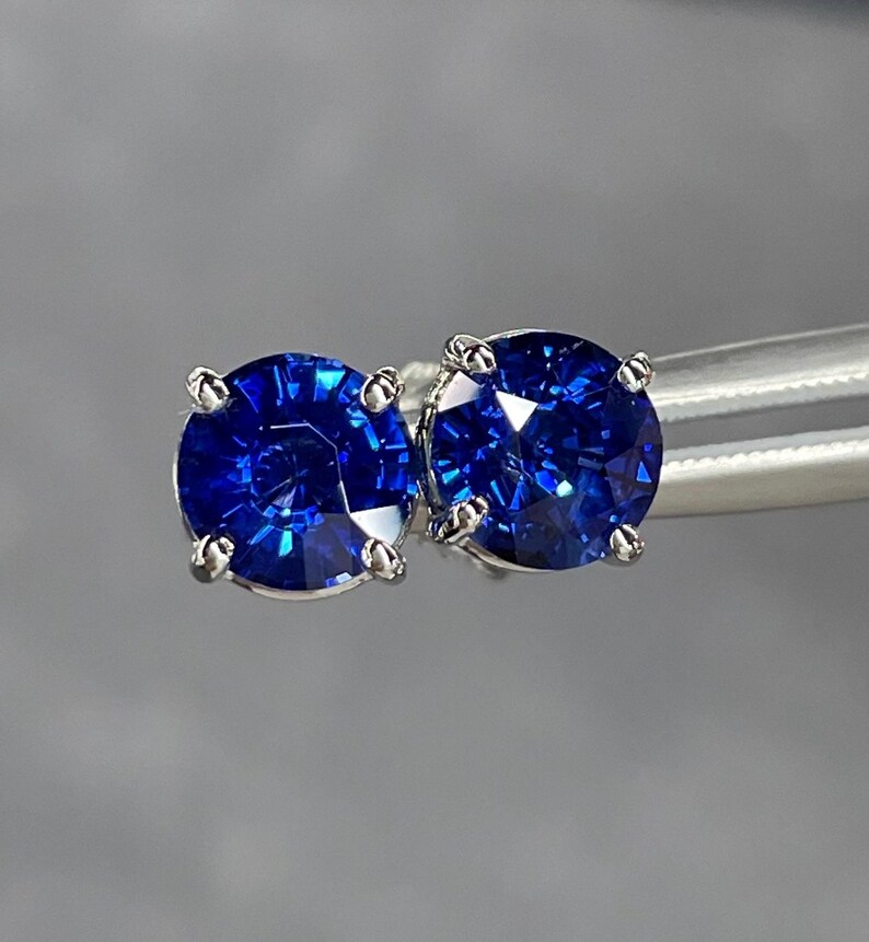 Pode incluir: Um par de brincos de press&atilde;o de safira azul redondos com corte brilhante. Cada brinco apresenta uma gema azul profunda e vibrante em uma crava&ccedil;&atilde;o de garras prateada. Os brincos s&atilde;o exibidos em um fundo cinza suave.