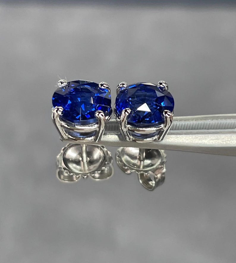 Pode incluir: Um par de brincos de press&atilde;o com pedras preciosas azuis profundas, redondas e brilhantes, engastadas em metal prateado. Os brincos s&atilde;o exibidos em uma superf&iacute;cie reflexiva, mostrando suas facetas e design. As pedras s&atilde;o o foco principal.