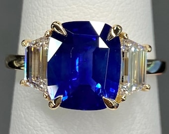 Pode incluir: Um anel de ouro com uma grande pedra preciosa azul vibrante, ladeada por duas pedras retangulares claras menores. A cravação do anel apresenta detalhes intrincados em ouro, criando um design luxuoso e atraente.