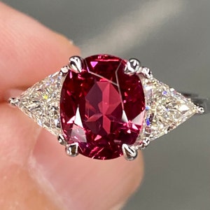 May include: A ring featuring a large oval red gemstone, flanked by two smaller triangular diamonds.