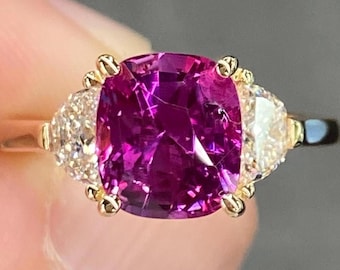 May include: A gold ring with a large, cushion-cut, magenta gemstone at its centre, with two smaller, triangular, clear gemstones on either side. The ring's band is plain, polished gold.