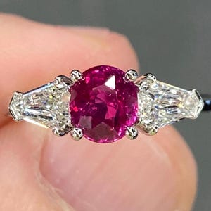 May include: A platinum ring featuring a vibrant, oval-cut ruby center stone flanked by two baguette-cut diamonds. The ruby is a deep pinkish-red color, and the diamonds sparkle brightly. The ring is a classic three-stone design.
