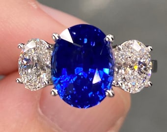 May include: A three-stone ring featuring a large oval-shaped blue sapphire flanked by two round brilliant-cut diamonds.