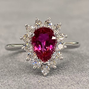 Puede incluir: Un anillo de metal blanco con una piedra preciosa rosa en forma de pera y un halo de pequeñas piedras preciosas transparentes.