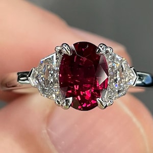 May include: A silver ring featuring a large, oval-cut, deep red gemstone as the centerpiece, flanked by two smaller, triangular, clear gemstones. The ring's band is a polished silver.