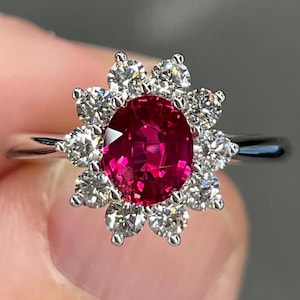 May include: A silver ring featuring a vibrant, oval-cut ruby center stone, surrounded by a halo of sparkling, round-cut diamonds. The ring's design resembles a flower, with the diamonds forming the petals.