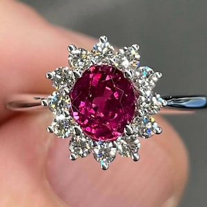 May include: A close-up of a silver ring featuring a vibrant, round, deep pink gemstone at the center, surrounded by a halo of smaller, sparkling white gemstones. The ring's band is thin and polished.