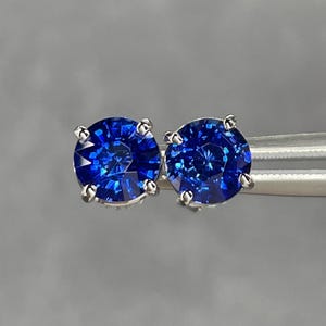 Pode incluir: Um par de brincos de press&atilde;o com pedras preciosas azuis redondas e lapidadas em brilhante. As pedras s&atilde;o engastadas em metal prateado com quatro garras. Os brincos s&atilde;o exibidos sobre um fundo cinza.