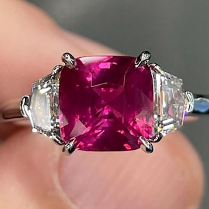 May include: A close-up of a silver ring featuring a large, vibrant, cushion-cut ruby center stone. Two smaller, rectangular, clear stones flank the ruby. The ring is set with four prongs.