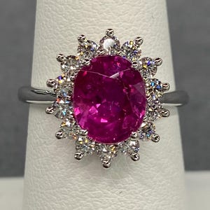 May include: A silver ring with a large, oval-cut, bright pink gemstone encircled by a halo of smaller, clear, round-cut diamonds. The ring's design is elegant, displaying a classic gemstone setting, suitable for special occasions.