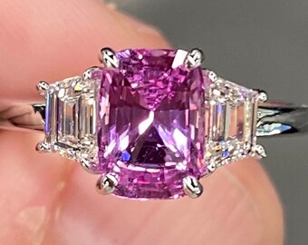 Pode incluir: Um anel de noivado de safira rosa com dois diamantes de corte baguette de cada lado da pedra central. O anel é engastado em ouro branco.