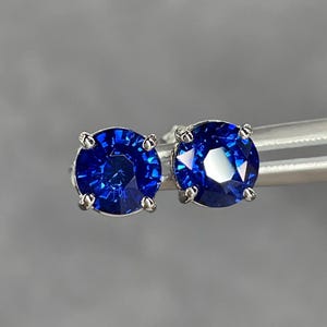 Pode incluir: Um par de brincos de press&atilde;o com pedras preciosas redondas azuis profundas. As pedras s&atilde;o engastadas em metal prateado com quatro garras. Os brincos s&atilde;o exibidos em um fundo cinza neutro.