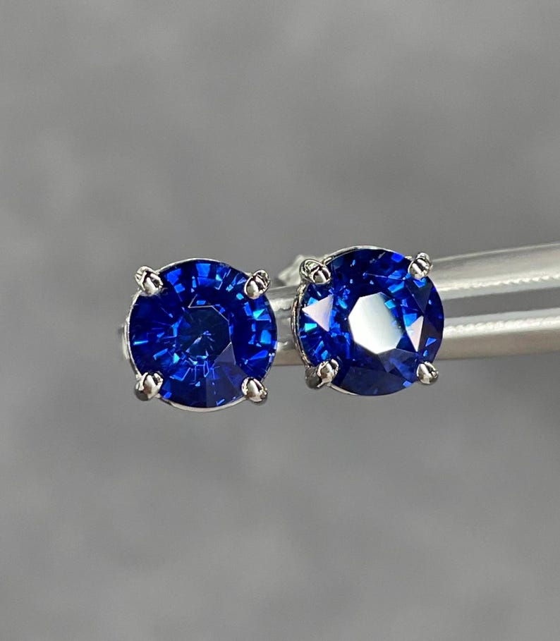 Pode incluir: Um par de brincos de press&atilde;o com pedras preciosas redondas azuis profundas. As pedras s&atilde;o engastadas em metal prateado com quatro garras. Os brincos s&atilde;o exibidos em um fundo cinza neutro.