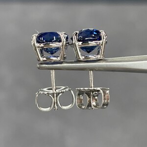 Pode incluir: Um par de brincos de press&atilde;o com pedras preciosas azuis escuras em metal prateado. Os brincos s&atilde;o segurados por uma ferramenta prateada, destacando o design intrincado das costas dos brincos. As pedras t&ecirc;m um corte redondo.