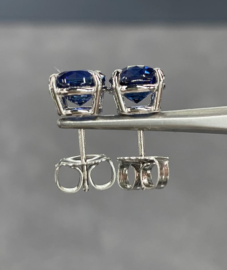 Pode incluir: Um par de brincos de press&atilde;o com pedras preciosas azuis escuras em metal prateado. Os brincos s&atilde;o segurados por uma ferramenta prateada, destacando o design intrincado das costas dos brincos. As pedras t&ecirc;m um corte redondo.