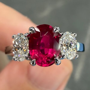 May include: A three-stone ring featuring a large oval ruby center stone flanked by two oval-cut diamonds. The ring has a silver band and claw prongs securing the gemstones. The ruby is a deep red color, and the diamonds sparkle brightly.
