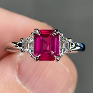 May include: A silver ring featuring a large, rectangular, vibrant red gemstone as the centerpiece, flanked by two smaller, clear, rectangular gemstones. The ring's band is a polished silver, and the setting has intricate details.