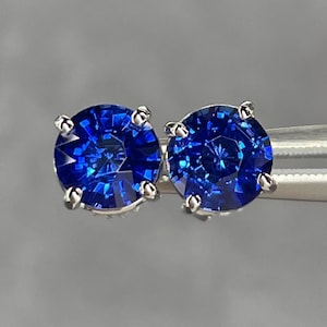 Pode incluir: Um par de brincos de bot&atilde;o redondos de pedras preciosas azuis profundas com corte brilhante. Cada brinco apresenta uma pedra azul vibrante em uma crava&ccedil;&atilde;o prateada de quatro garras. Os brincos s&atilde;o exibidos em um fundo cinza neutro.