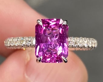 Pode incluir: Um anel de noivado de safira rosa com uma pedra de corte esmeralda quadrada engastada em uma banda de ouro branco com um halo de diamantes.