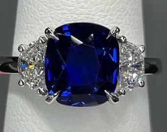 May include: A three-stone ring featuring a large, cushion-cut deep blue sapphire centre stone set in polished silver-toned metal. The centre sapphire is flanked by two smaller, half-moon-shaped clear white diamonds on each side in a prong setting.