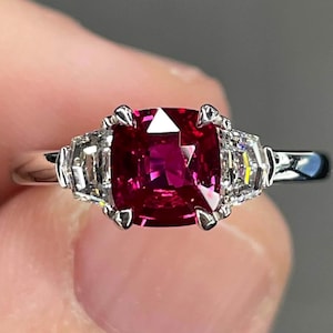 May include: A close-up of a platinum ring featuring a large, square-cut, deep red gemstone as the centerpiece. Flanking the central stone are two smaller, rectangular, clear gemstones. The ring's band is a polished silver color.
