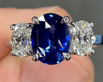 May include: A three-stone ring featuring a large, oval-cut, deep blue sapphire center stone flanked by two smaller, oval-cut, clear diamond side stones. The ring is set in a silver metal band.