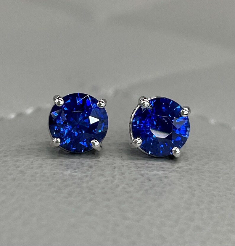 Pode incluir: Um par de brincos de press&atilde;o com pedras preciosas redondas, lapidadas em brilhante, azul profundo. As pedras s&atilde;o engastadas em metal prateado com quatro garras. Os brincos s&atilde;o exibidos em uma superf&iacute;cie cinza.