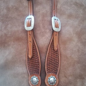 May include: A pair of brown leather horse headstalls with silver buckles and conchos. The headstalls have a woven pattern and are stitched with white thread.