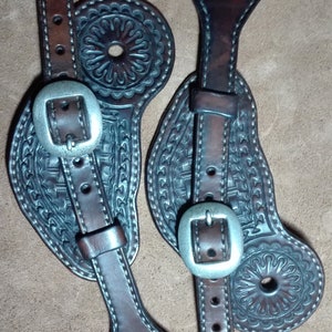 May include: Two brown leather boot straps with silver buckles and intricate tooled designs. The straps have a loop at the top and a hole at the bottom.
