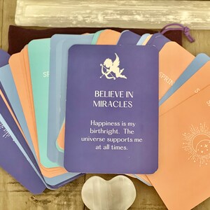 May include: A deck of oracle cards with a central purple card bearing the text "BELIEVE IN MIRACLES". The cards are in shades of purple, blue, and peach. A heart-shaped crystal and a small pouch are also present.