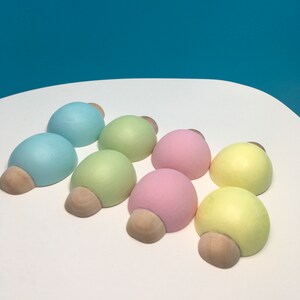 May include: Eight pastel-coloured wooden decorative objects, each with a rounded top and a light brown, half-circle base. The colours include light blue, green, pink, and yellow. The objects are arranged on a white surface.