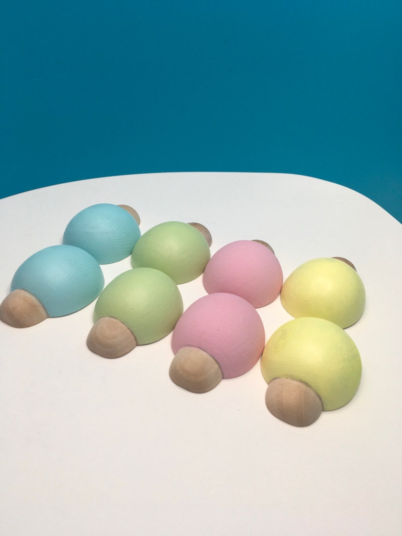 May include: Eight pastel-coloured wooden decorative objects, each with a rounded top and a light brown, half-circle base. The colours include light blue, green, pink, and yellow. The objects are arranged on a white surface.