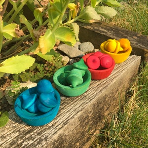 May include: A selection of brightly coloured wooden toy fruits and vegetables in small bowls. The set includes blue, green, red, and yellow items, arranged on a weathered wooden surface. The toys are designed to resemble various types of produce.