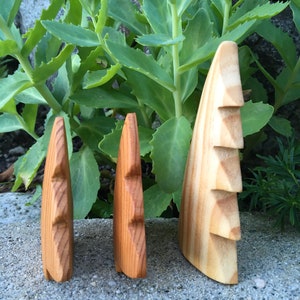May include: Three wooden sculptures of stylized faces, each with a unique size and wood grain. The sculptures are displayed outdoors against a backdrop of green foliage and a grey stone surface. The wood tones range from light to medium brown.