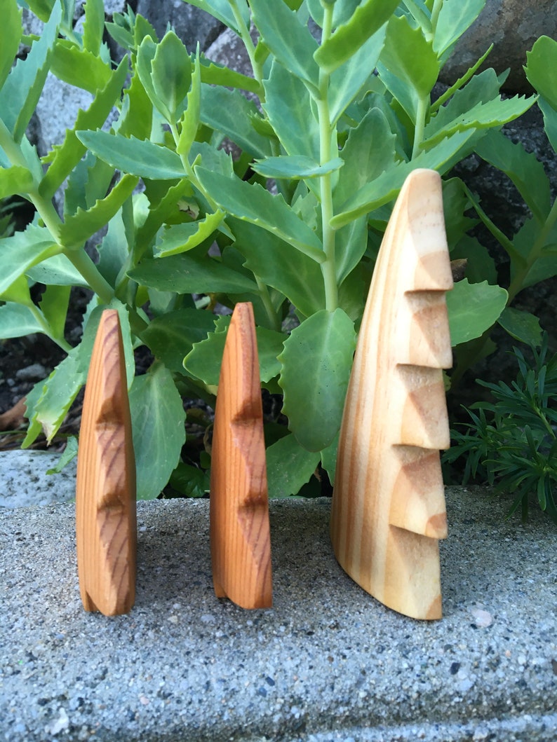 May include: Three wooden sculptures of stylized faces, each with a unique size and wood grain. The sculptures are displayed outdoors against a backdrop of green foliage and a grey stone surface. The wood tones range from light to medium brown.