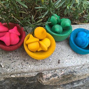 May include: Four small wooden bowls, each filled with colourful wooden acorns. The acorns are painted in bright shades of red, yellow, green, and blue. The bowls sit on a concrete surface with greenery in the background.