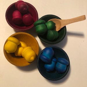 May include: Four small wooden bowls, each filled with colourful wooden acorns. The bowls are in shades of red, green, yellow, and blue. A small wooden spoon rests in one of the green bowls.