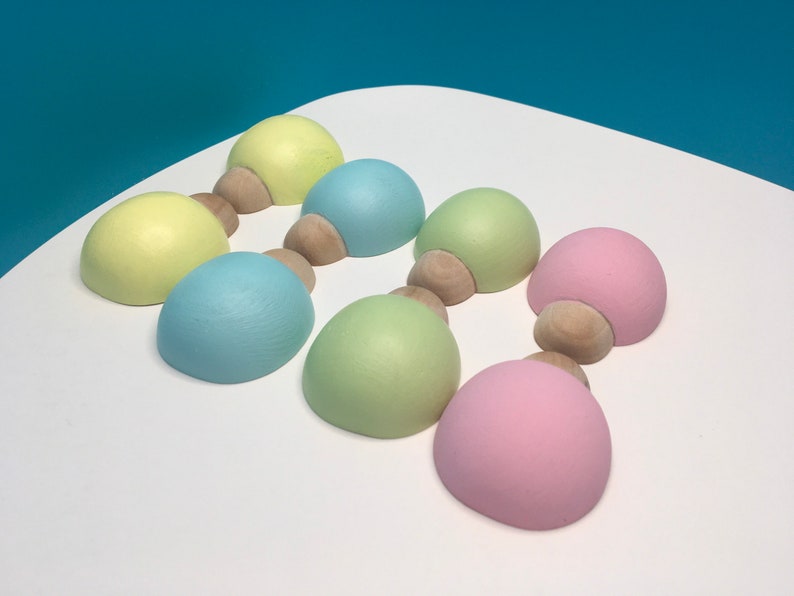 May include: Seven pastel-coloured wooden decorative objects. Each object has a rounded top in shades of yellow, blue, green, and pink, connected to a small, natural wood base. The objects are arranged on a white surface.