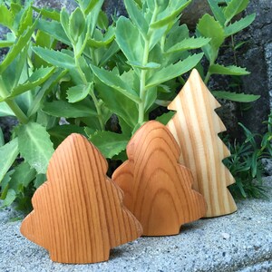 May include: Three wooden Christmas tree ornaments. Two are a warm brown colour with a rounded shape, and the third is a lighter wood colour with a traditional tree shape. The ornaments are displayed on a grey stone surface with green foliage in the background.