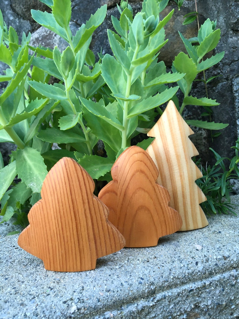 May include: Three wooden Christmas tree ornaments. Two are a warm brown colour with a rounded shape, and the third is a lighter wood colour with a traditional tree shape. The ornaments are displayed on a grey stone surface with green foliage in the background.